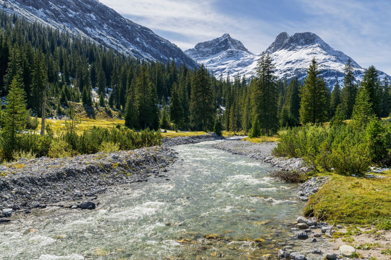 Ein klarer Fluss in den Alpen, umgeben von schneebedeckten Bergen, erinnert an die natürliche Schönheit auf dem Weg zur Besteigung Kilimandscharo.