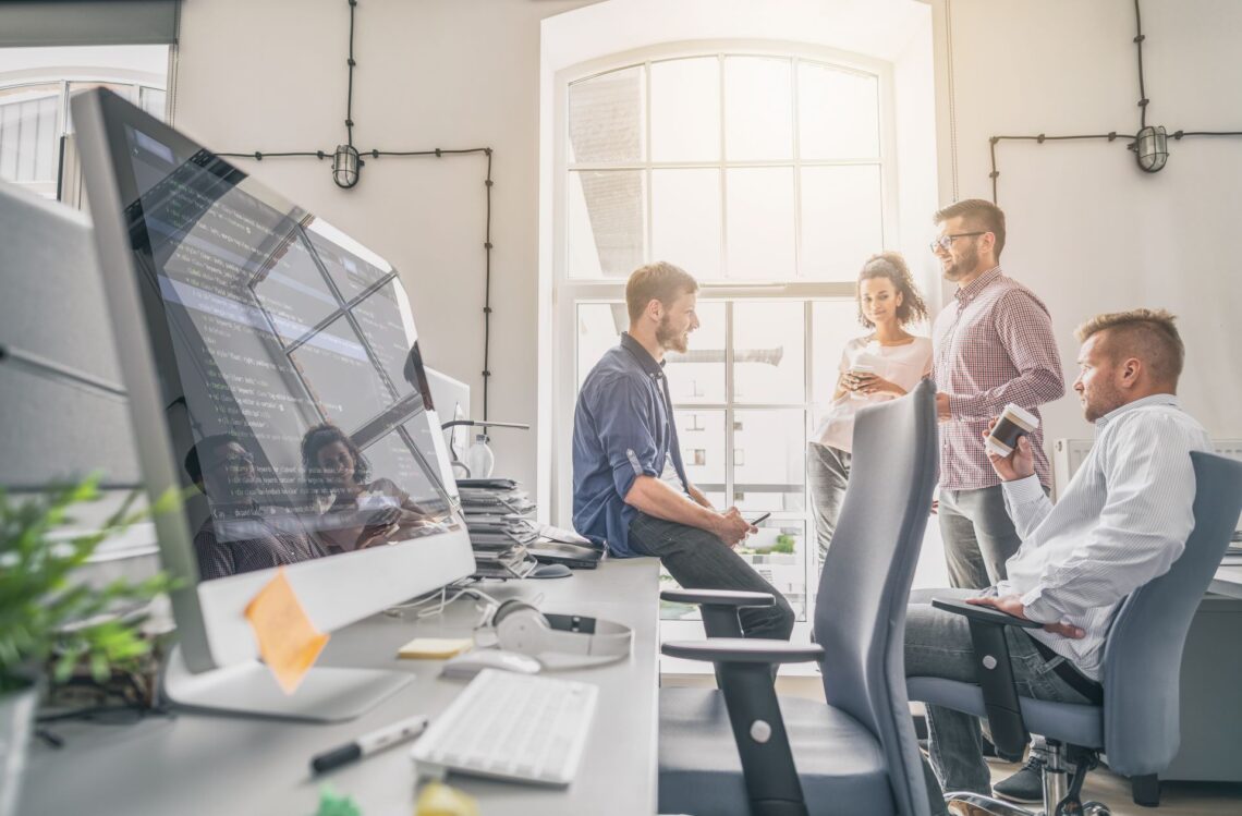 Modernes Agentur-Büro mit Teammeeting – Junge Fachkräfte diskutieren in einem lichtdurchfluteten Büro mit großem Computerbildschirm.